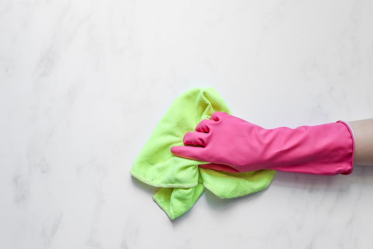 about-01 Pink rubber glove cleaning a surface with a green cloth on a white background.
